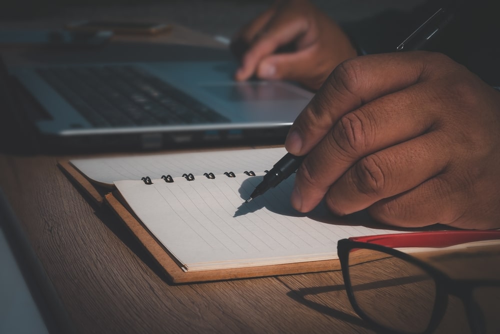 Person Working On A Computer And Writing In A Notebook